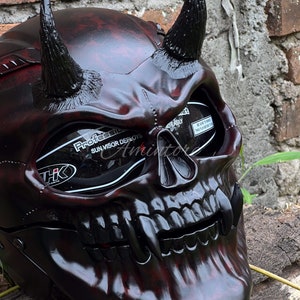 K&ouml;nnte beinhalten: Ein schwarzer Motorradhelm mit Totenkopf-Design und roten Akzenten. Der Helm hat H&ouml;rner und ein get&ouml;ntes Visier. Der Text "Professional Sun Visor Deploys" ist auf dem Visier sichtbar.