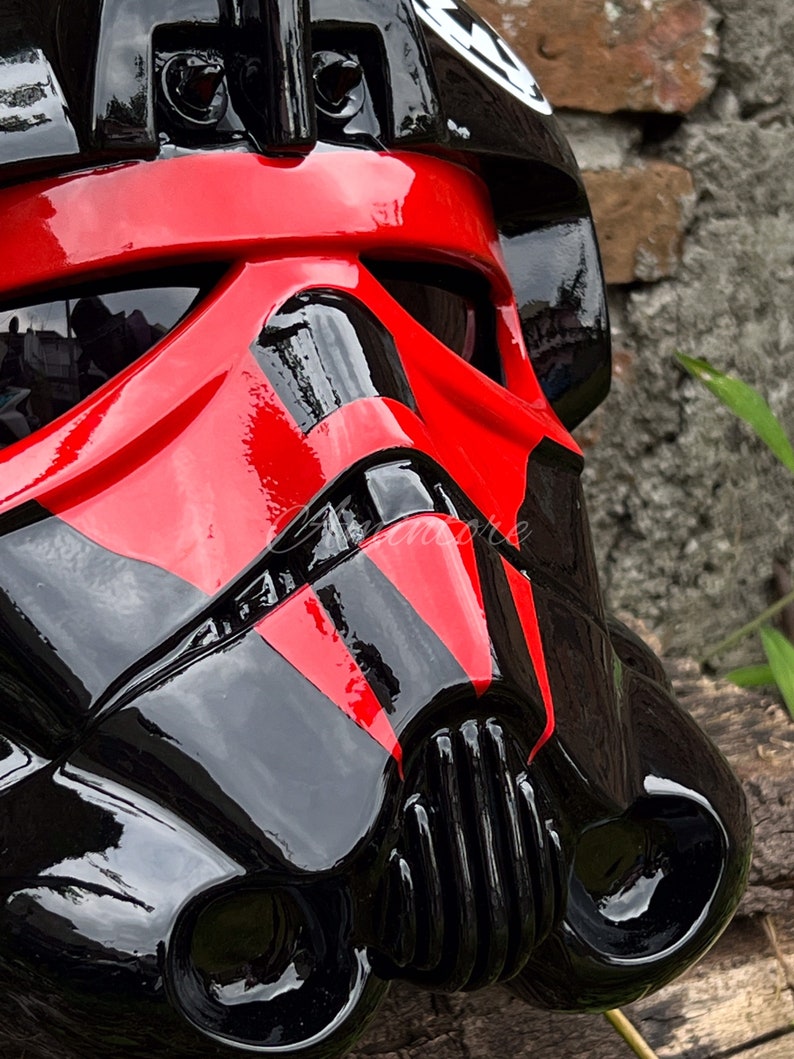 May include: Close-up of a glossy black and red helmet. The helmet features a distinctive design with red accents across the face and a black visor. A white emblem is visible on the top of the helmet. The helmet is likely a costume or collectible item.