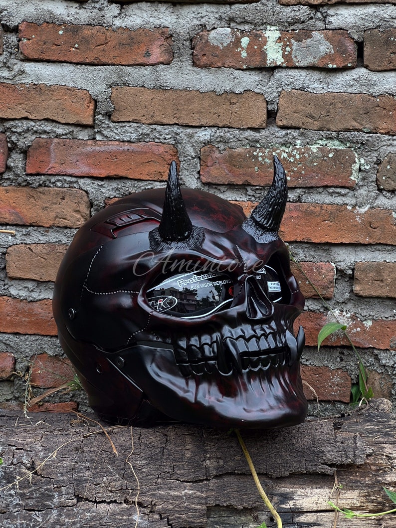 K&ouml;nnte beinhalten: Ein schwarzer und roter Motorradhelm mit Totenkopf-Design und H&ouml;rnern. Der Helm hat ein klares Visier mit dem aufgedruckten Text "Professional Visor Deploys".