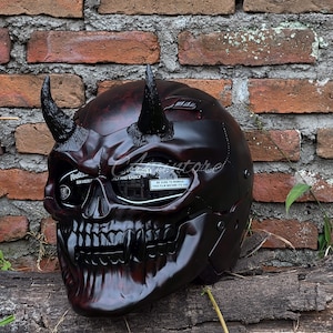 K&ouml;nnte beinhalten: Ein schwarzer und roter Motorradhelm mit Totenkopf-Design und H&ouml;rnern. Der Helm hat ein klares Visier mit einer Schutzfolie, auf der "BE SURE TO REMOVE THIS FILM BEFORE ITS USE" steht.