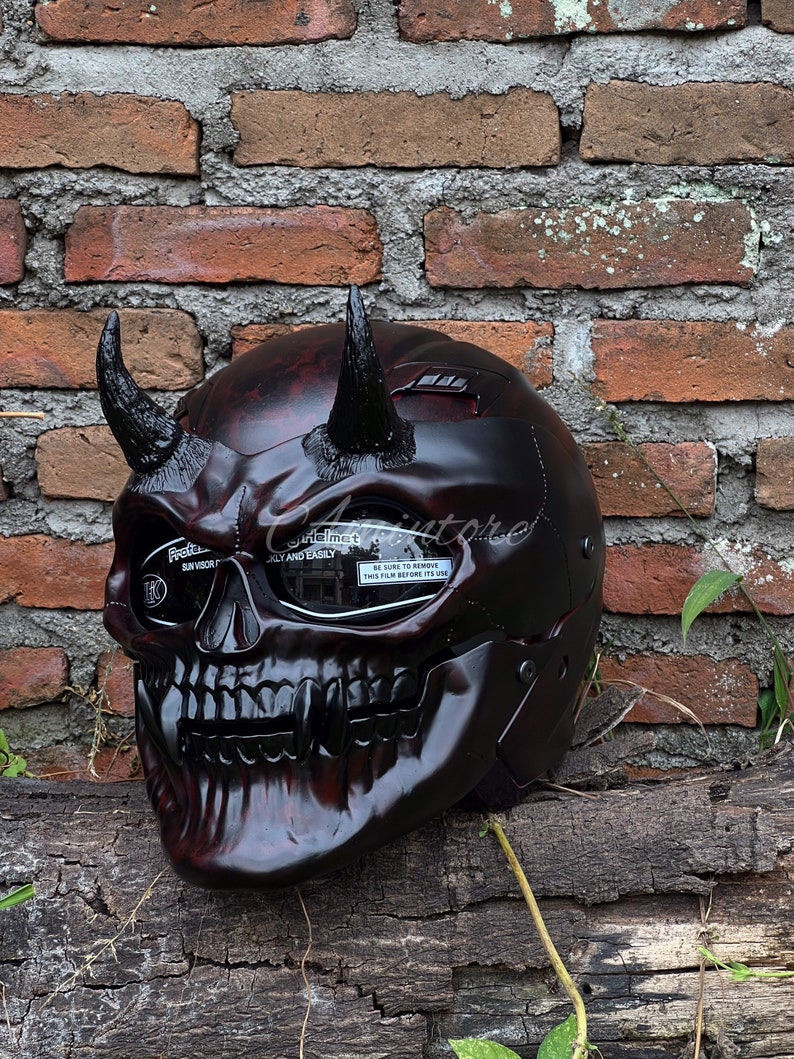 K&ouml;nnte beinhalten: Ein schwarzer und roter Motorradhelm mit Totenkopf-Design und H&ouml;rnern. Der Helm hat ein klares Visier mit einer Schutzfolie, auf der "BE SURE TO REMOVE THIS FILM BEFORE ITS USE" steht.
