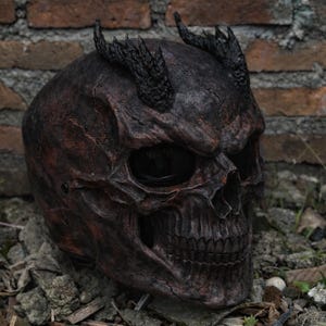 May include: A detailed, dark brown skull helmet with textured, cracked surfaces and black horns. The helmet has a menacing appearance, with large eye sockets and a detailed jawline. The background features a brick wall and foliage.
