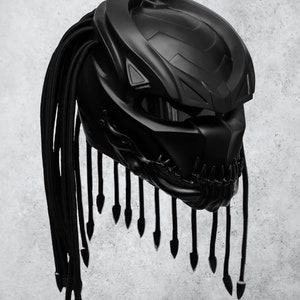 Op de afbeelding: Een zwarte motorhelm met een predator-achtig ontwerp en een lange zwarte vlecht. De helm heeft een vizier en een getextureerd oppervlak.