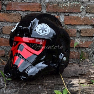 May include: A glossy black helmet with red accents, featuring white Imperial insignias. The helmet is a replica of a Star Wars TIE fighter pilot helmet, with a detailed design and a smooth, reflective surface.