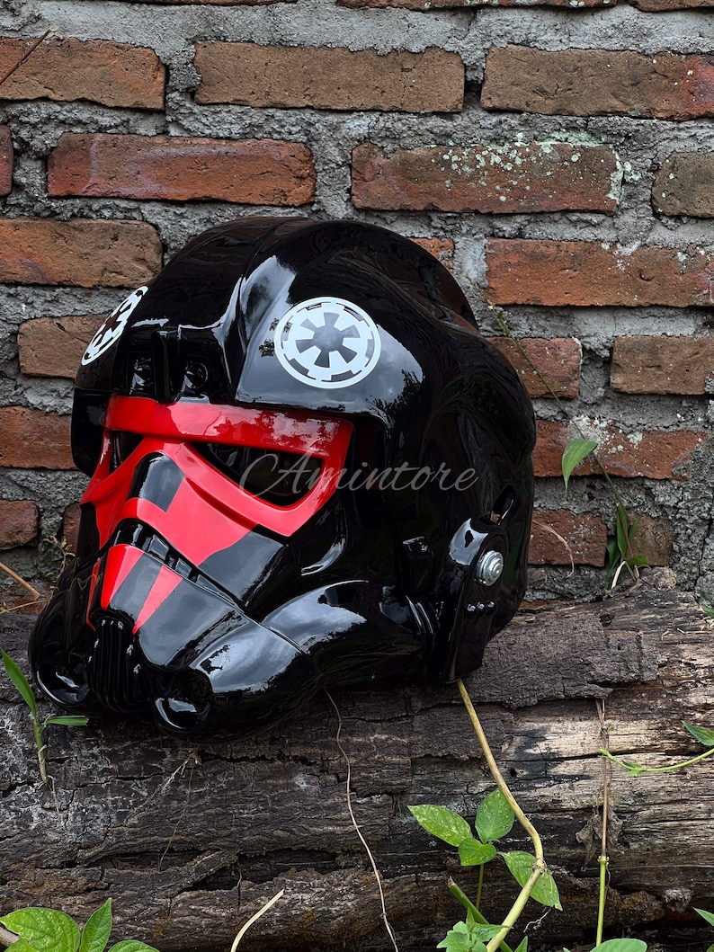 May include: A glossy black helmet with red accents, featuring white Imperial insignias. The helmet is a replica of a Star Wars TIE fighter pilot helmet, with a detailed design and a smooth, reflective surface.
