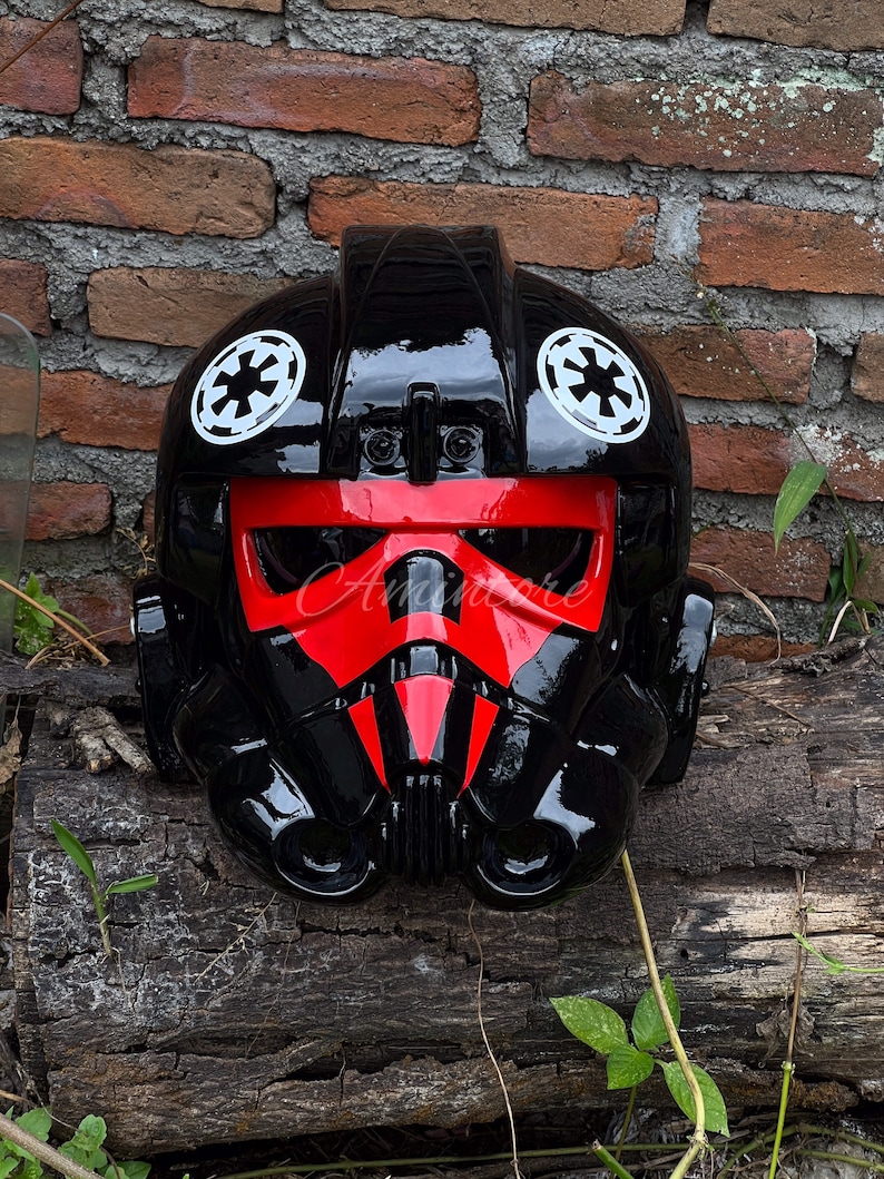 May include: A black and red TIE fighter pilot helmet with white Imperial insignias. The helmet has a glossy finish and a distinctive red face mask. A Star Wars themed collectible.