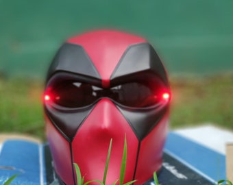 May include: A red and black Deadpool mask with a black visor and red glowing eyes. The mask is sitting on a blue and white surface with green grass in the foreground.