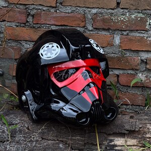 May include: A glossy black helmet with red accents, featuring white circular emblems. The helmet has a distinctive visor and ventilation details, resting on a weathered wooden surface against a brick wall.