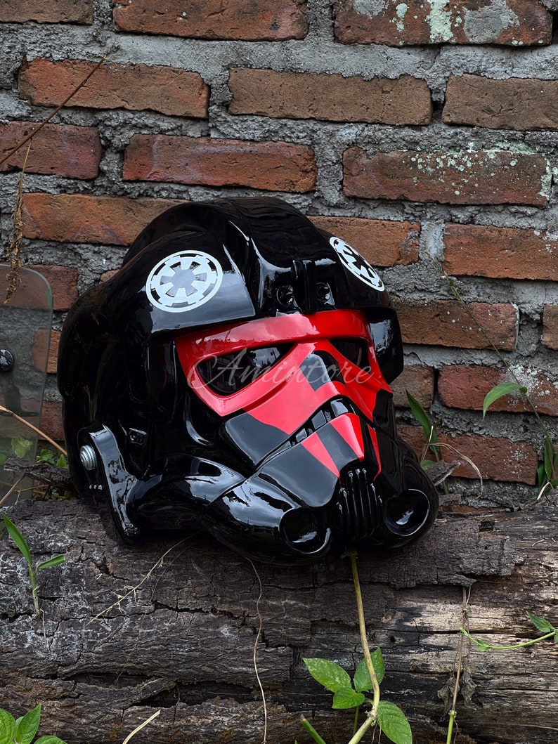 May include: A glossy black helmet with red accents, featuring white circular emblems. The helmet has a distinctive visor and ventilation details, resting on a weathered wooden surface against a brick wall.