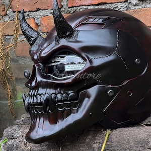 K&ouml;nnte beinhalten: Ein schwarzer und roter Motorradhelm mit Totenkopf-Design und H&ouml;rnern. Der Helm hat ein klares Visier und einen schwarzen Riemen.