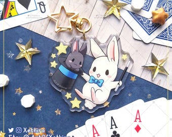 May include: A clear acrylic charm featuring a white rabbit holding a magic wand and a black rabbit wearing a blue and white top hat. The charm is surrounded by gold stars and is attached to a gold key chain.