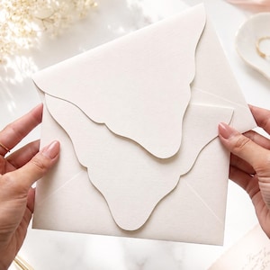 Può includere: Una busta color crema caratterizzata da un design con patta smerlata. La carta presenta una finitura testurizzata visibile e la busta è realizzata con un bordo decorativo a punta che si sovrappone al corpo principale della cancelleria.