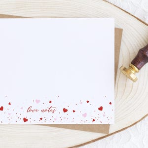 May include: A white card with the words "love notes" in cursive, and a border of red and pink hearts. The card is on a wooden slice with a brown envelope behind it.