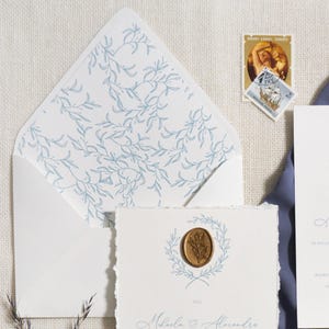May include: Wedding stationery set with a white invitation card featuring a blue floral wreath and a gold wax seal. Includes a matching envelope with a blue leaf pattern, a blue ribbon, and a postage stamp.