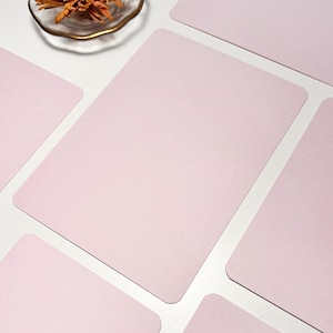 May include: Pale pink rectangular placemats with rounded corners, arranged on a white surface. A small glass dish with dried flowers is in the upper left. The placemats are a solid colour. Each measures approximately 30 cm x 20 cm.