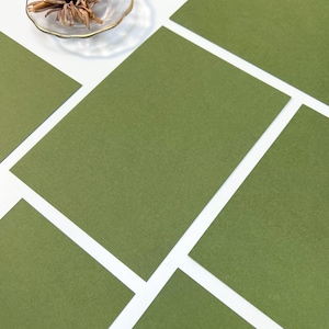 May include: Olive green stationery cards arranged on a white surface. A dried flower and a decorative stamp are also present. The cards are rectangular and have a textured appearance. The arrangement creates a visually appealing layout.