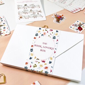 Könnte beinhalten: Eine weiße Box mit einem floralen Band mit der Aufschrift "THE BOOK LOVER'S BOX". Das Bild enthält ein Lesezeichen, Aufkleber und einen Lesetracker. Die Artikel haben ein florales Design mit rosa, gelben und grünen Akzenten.
