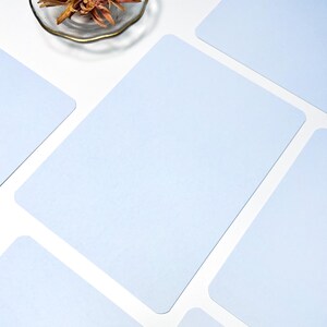 May include: Several pale blue rectangular placemats with rounded corners are arranged on a white surface. A small glass dish with dried flowers is in the upper left corner.