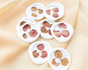 Peut inclure: Sceaux de cire blanche avec des fleurs roses séchées pressées dans la cire. Les sceaux sont éparpillés sur un fond de tissu pêche clair. Un tampon à cire en bois et en or est également visible sur l'image.