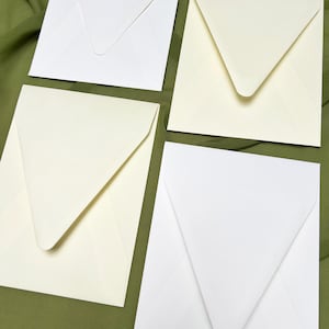Puede incluir: Cuatro sobres cuadrados en tonos blanco y crema sobre tela verde. Cada sobre presenta una solapa puntiaguda clásica. El papel tiene un acabado liso, ideal para papelería o invitaciones. La disposición enfatiza el diseño y la presentación.
