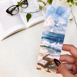 May include: A watercolour bookmark depicting a seascape with a sailboat, waves, and a sandy beach. The bookmark is held near an open book and black-framed glasses. Green leaves and a white background complete the scene.