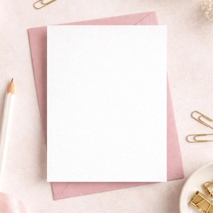 May include: A blank white card on a pink envelope, with a white pencil, gold paper clips, and a pink ribbon. The stationery is arranged on a light pink surface, with a small white dish filled with gold paper clips and a sprig of white flowers.