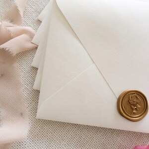 May include: White envelope with a gold wax seal featuring a rose design. The envelope is on a textured white surface with a pink ribbon.