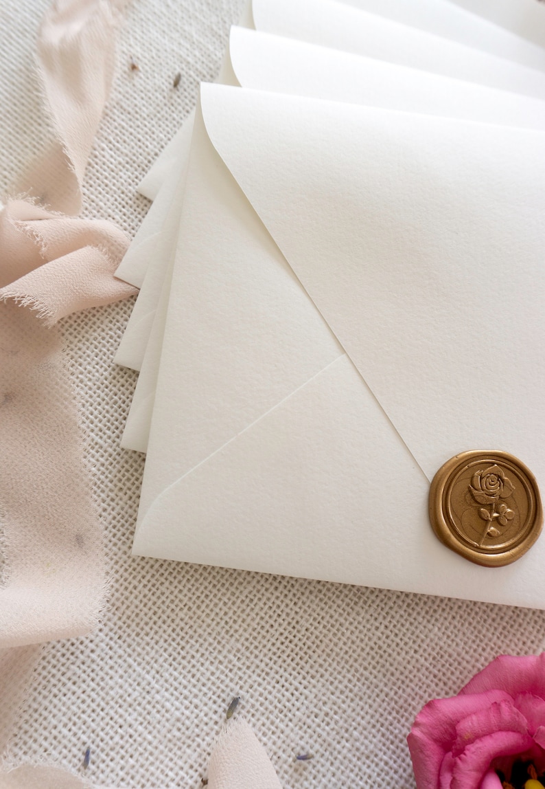 May include: White envelope with a gold wax seal featuring a rose design. The envelope is on a textured white surface with a pink ribbon.
