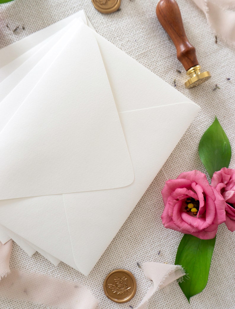 May include: White envelopes with a pink flower and gold wax seals on a textured fabric background.