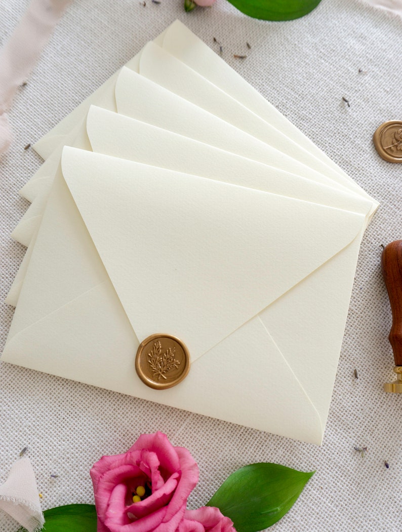 May include: A stack of ivory envelopes with a gold wax seal featuring a floral design. The envelopes are on a white textured surface with pink flowers and green leaves.