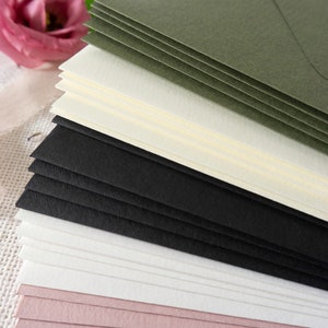 K&ouml;nnte beinhalten: Ein Stapel Umschl&auml;ge in verschiedenen Farben, darunter Schwarz, Wei&szlig;, Creme, Rosa und Gr&uuml;n. Die Umschl&auml;ge sind versetzt angeordnet, wobei die gr&uuml;nen Umschl&auml;ge oben liegen.