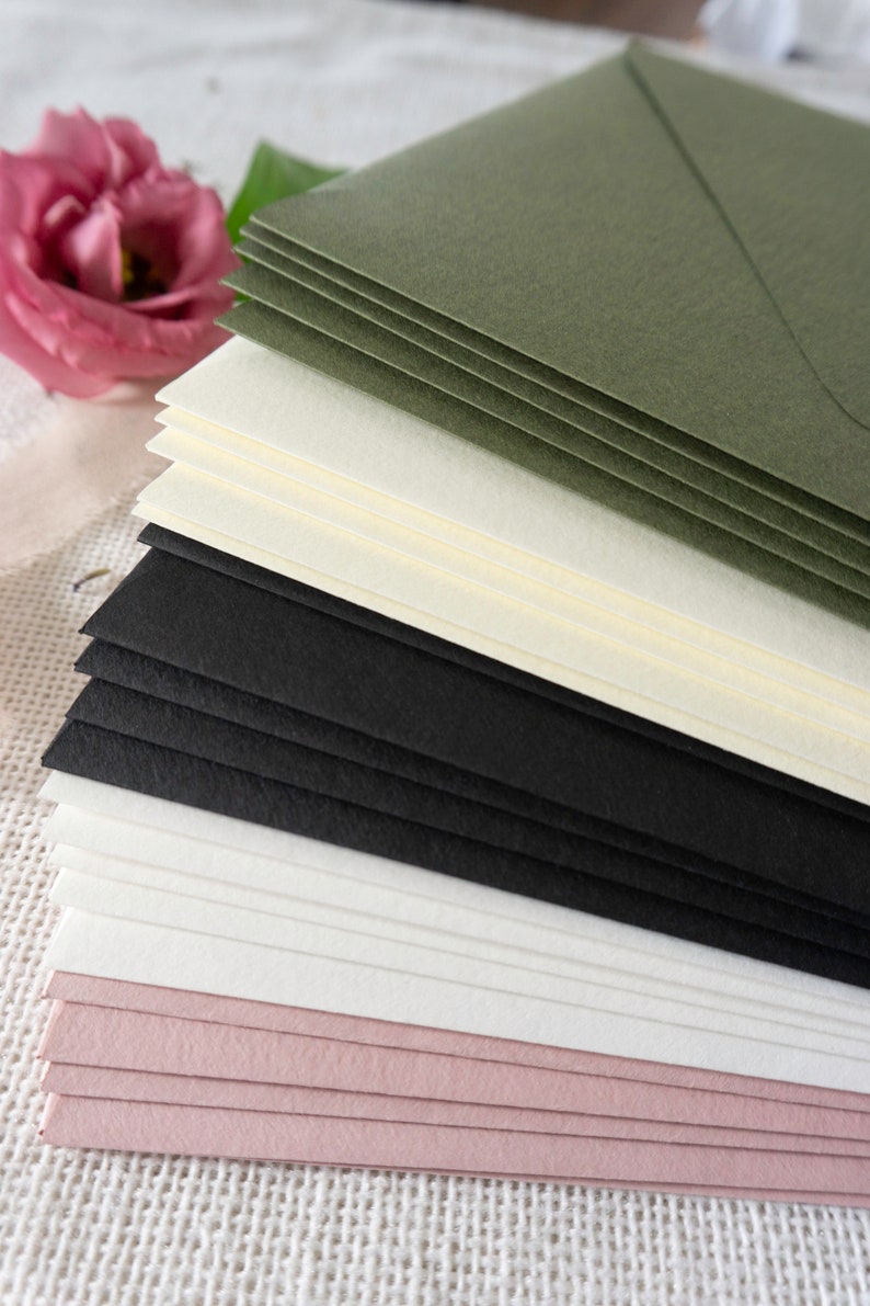 K&ouml;nnte beinhalten: Ein Stapel Umschl&auml;ge in verschiedenen Farben, darunter Schwarz, Wei&szlig;, Creme, Rosa und Gr&uuml;n. Die Umschl&auml;ge sind versetzt angeordnet, wobei die gr&uuml;nen Umschl&auml;ge oben liegen.