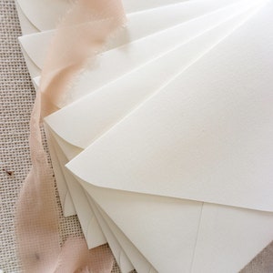 May include: A stack of ivory envelopes with rounded corners, arranged in a fan shape. The envelopes are on a textured beige fabric surface with a piece of peach-colored ribbon.
