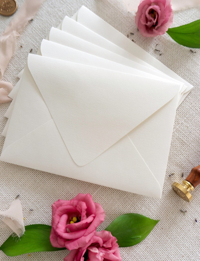 May include: A stack of white envelopes on a textured beige surface with pink flowers and a gold wax seal.
