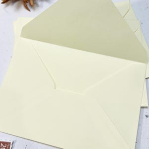 以下が含まれることがあります： クリーム色の封筒の束。1つは開いており、内側が明るくなっています。封筒は明るい色の表面に配置され、背景にはドライフラワーと切手が見えます。