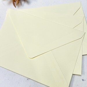 以下が含まれることがあります： クリーム色の封筒の束。封筒は長方形で、三角形のフラップが付いています。紙は滑らかな質感です。封筒はわずかに重なり合うように配置されており、上の封筒が他の封筒を部分的に覆っています。