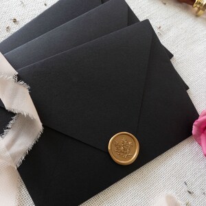 K&ouml;nnte beinhalten: Drei schwarze Umschl&auml;ge mit einem goldenen Siegel aus Wachs auf dem oberen Umschlag. Das Siegel aus Wachs hat ein florales Design.