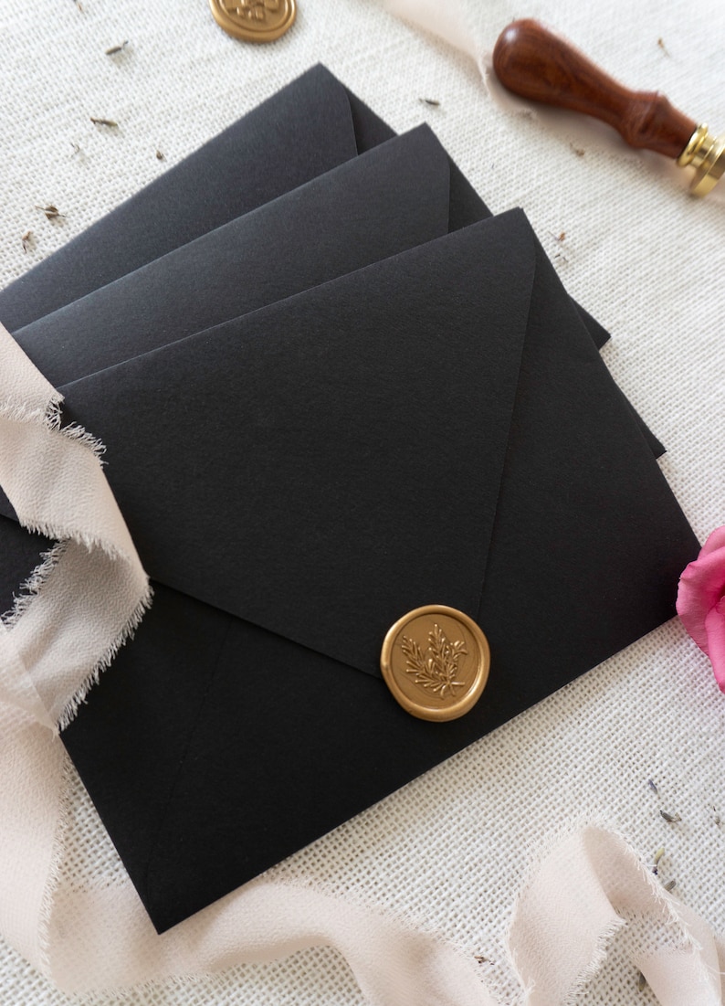 K&ouml;nnte beinhalten: Drei schwarze Umschl&auml;ge mit einem goldenen Siegel aus Wachs auf dem oberen Umschlag. Das Siegel aus Wachs hat ein florales Design.