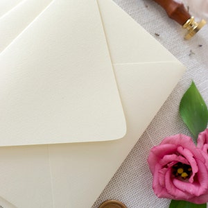 May include: A stack of ivory envelopes with a gold wax seal and a pink flower on a textured fabric background.