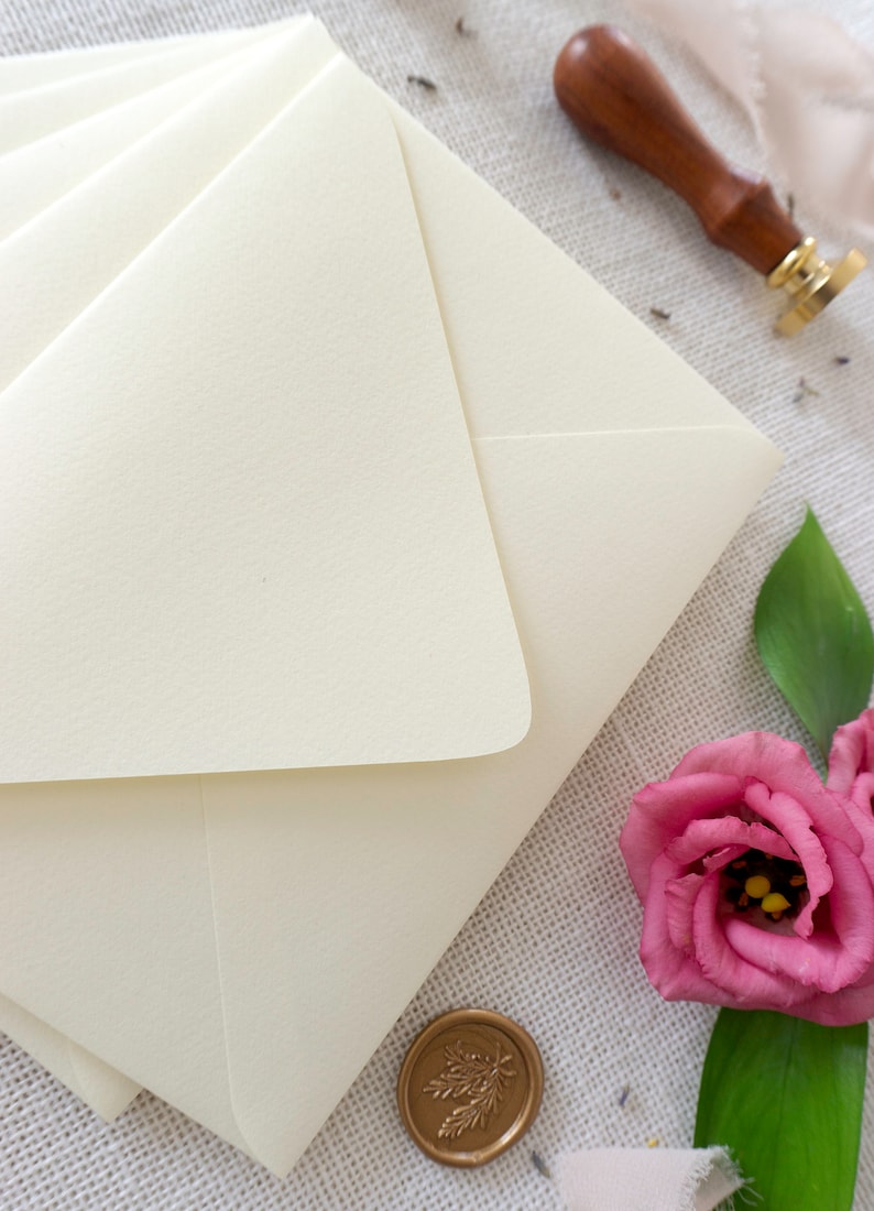 May include: A stack of ivory envelopes with a gold wax seal and a pink flower on a textured fabric background.