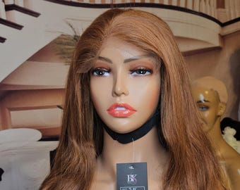 May include: A long, wavy, light brown human hair wig on a mannequin head. The wig is styled in loose waves and has a natural-looking parting. The wig is on a black stand and has a black label attached to it with the text "BK Beauty" and "13x4".