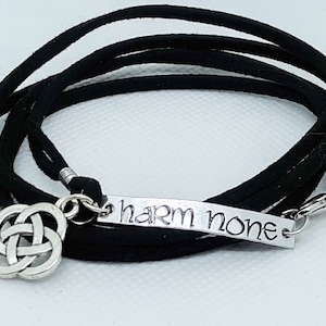 May include: Black wrap bracelet with a silver charm featuring a Celtic knot design and a silver tag with the words "harm none".