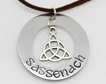 May include: Silver necklace with a round pendant featuring a Celtic knot design and the word "Sassenach" inscribed around the edge. The necklace is made of suede cord.