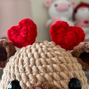 Puede incluir: Un juguete de peluche de ganchillo en forma de oso marr&oacute;n con grandes ojos negros. El oso lleva dos corazones de fieltro rojo en la cabeza.
