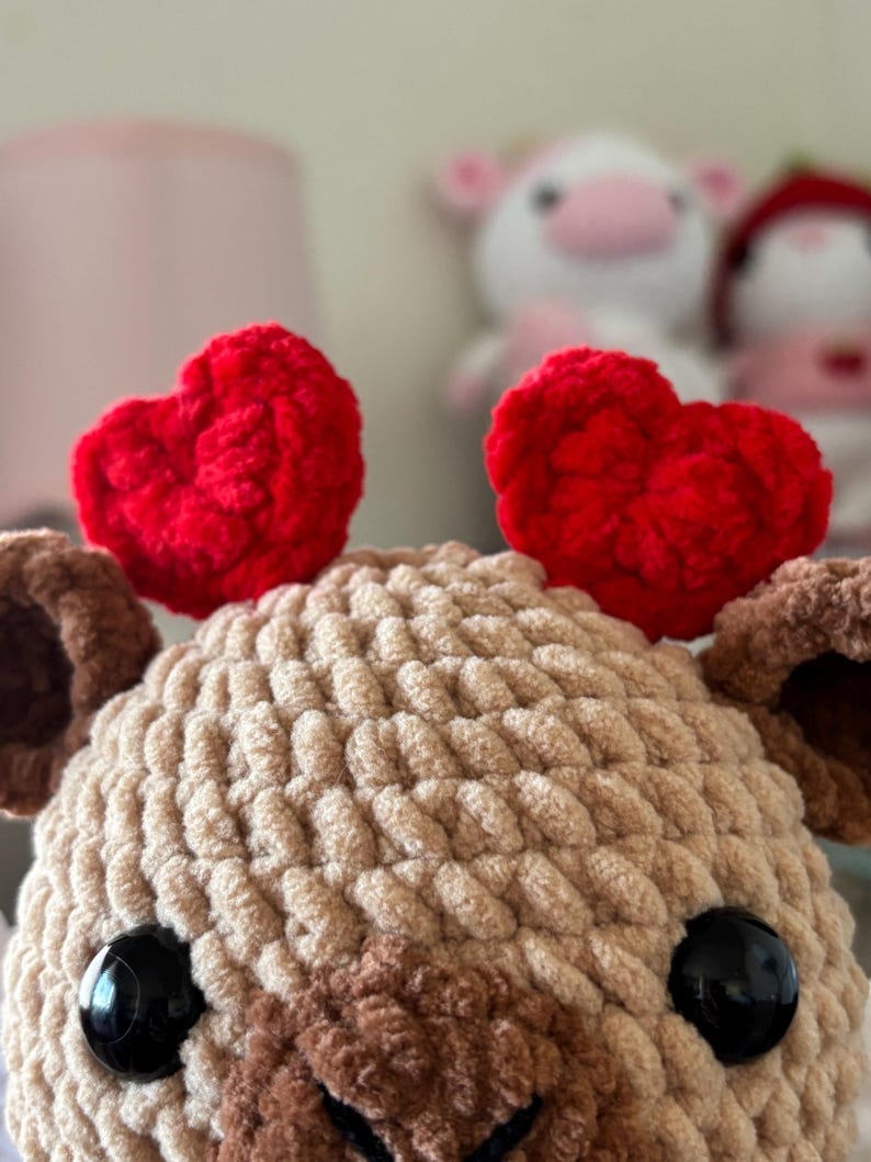 Puede incluir: Un juguete de peluche de ganchillo en forma de oso marr&oacute;n con grandes ojos negros. El oso lleva dos corazones de fieltro rojo en la cabeza.