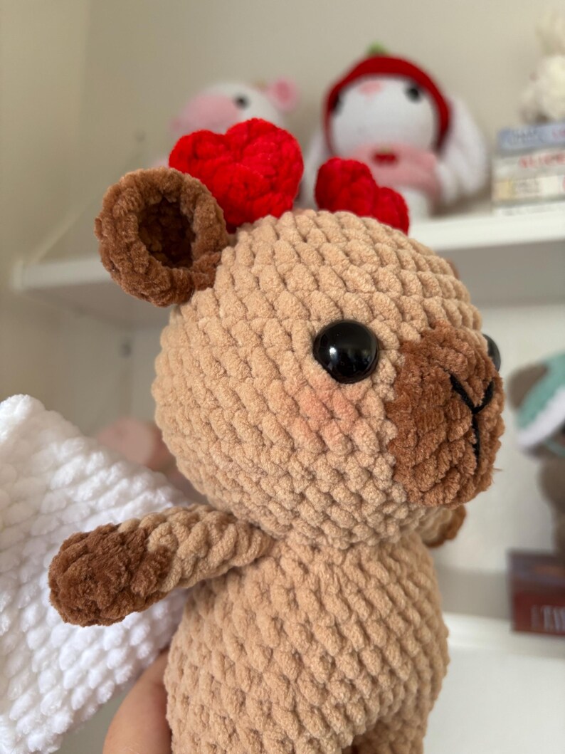 Puede incluir: Un juguete de peluche de color marr&oacute;n tejido a crochet con corazones de fieltro rojo en la cabeza y alas blancas tejidas a crochet. El juguete tiene ojos negros y una nariz marr&oacute;n.