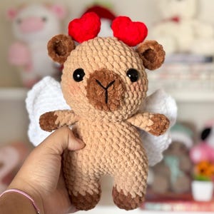 Puede incluir: Un juguete de peluche de ganchillo en forma de oso marr&oacute;n con alas blancas y orejas en forma de coraz&oacute;n rojo. El oso tiene una nariz marr&oacute;n y ojos negros.