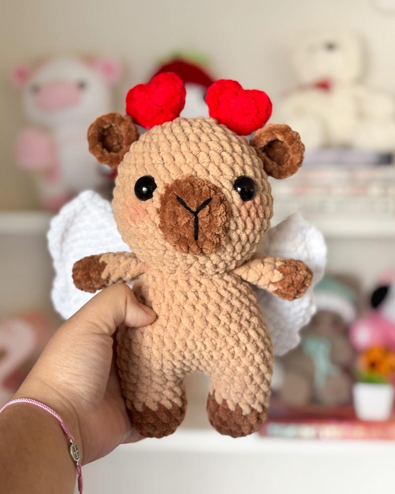 Puede incluir: Un juguete de peluche de ganchillo en forma de oso marr&oacute;n con alas blancas y orejas en forma de coraz&oacute;n rojo. El oso tiene una nariz marr&oacute;n y ojos negros.