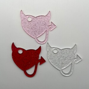 May include: Three glitter heart-shaped keychains with a devil tail design. The keychains are pink, red, and silver.