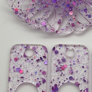 May include: Two clear rectangular keychains with a purple glitter embedded in the resin. Each keychain has a round hole in the center and a smaller hole at the top for attaching a ring.